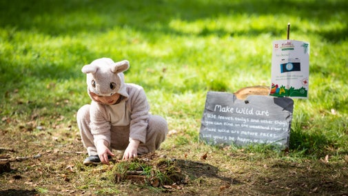 Small child in pale pink bunny outfit, making 'wild art' in a garden, with a slate 'wild art' sign behind